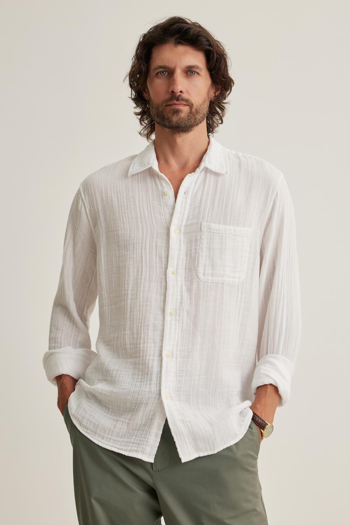 A man with medium-length hair and a beard wears the Velvet by Graham & Spencer ELTON COTTON GAUZE BUTTON-UP SHIRT, in white, paired with green pants while standing against a plain backdrop with his hands in his pockets.-38643274186945