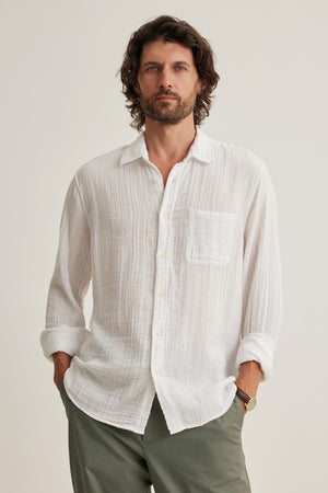 A man with medium-length hair and a beard wears the Velvet by Graham & Spencer ELTON COTTON GAUZE BUTTON-UP SHIRT, in white, paired with green pants while standing against a plain backdrop with his hands in his pockets.