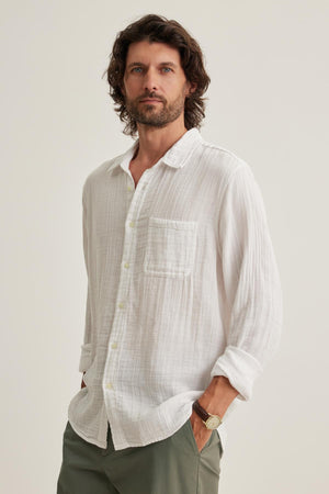 Against a neutral background, a man with wavy hair dons the ELTON COTTON GAUZE BUTTON-UP SHIRT by Velvet by Graham & Spencer, with his hands in his pockets.