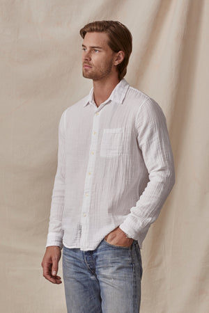 A man in a Velvet by Graham & Spencer Elton Cotton Gauze button-up shirt with double button cuffs and faded blue jeans, standing with his hand in his pocket against a neutral beige background.