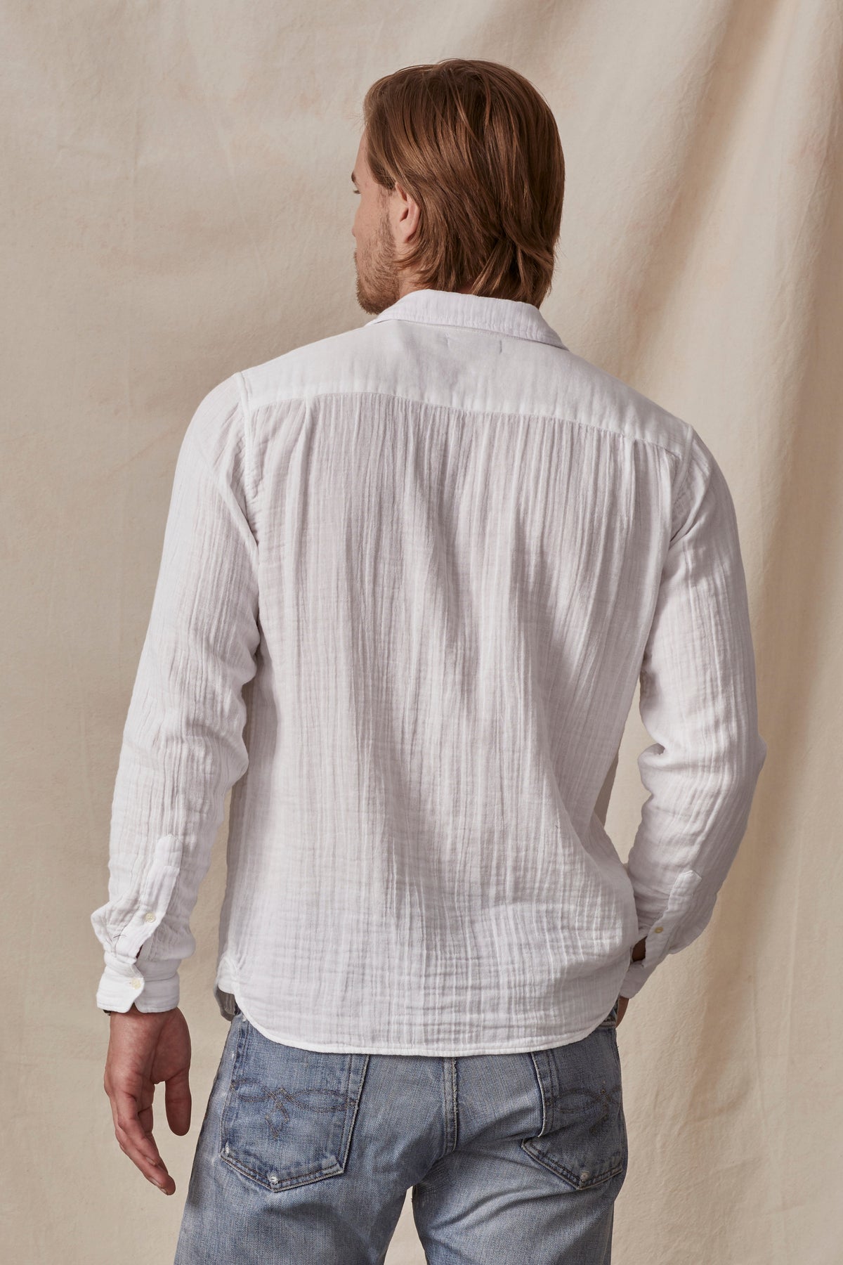  A man viewed from the back, wearing a Velvet by Graham & Spencer ELTON COTTON GAUZE BUTTON-UP SHIRT and blue jeans, against a neutral beige background. 