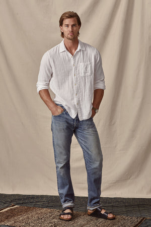 Man standing against a cream backdrop, wearing a Velvet by Graham & Spencer Elton Cotton Gauze Button-Up Shirt, blue jeans, and black sandals, with his left hand in his pocket.