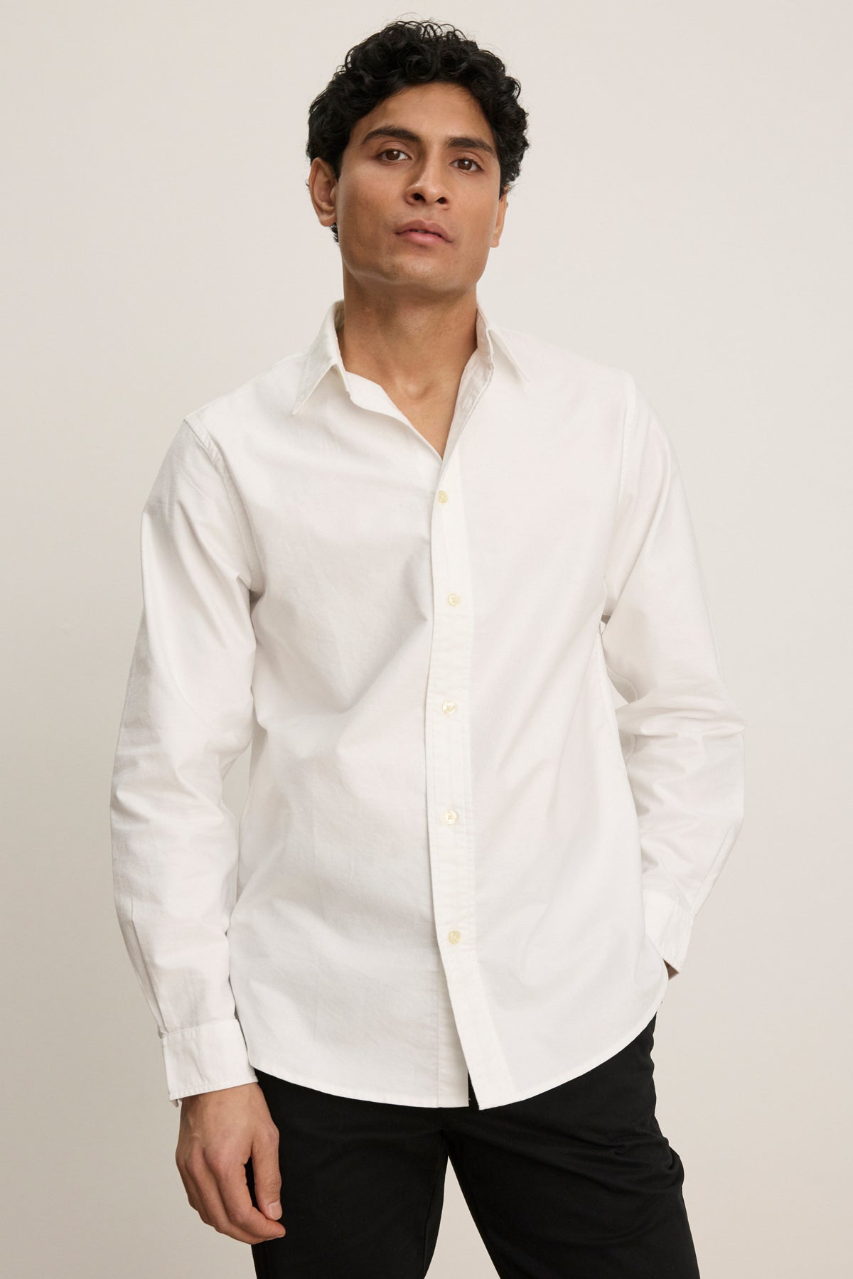 A man with short dark hair wearing the EMMETT BUTTON-DOWN SHIRT by Velvet by Graham & Spencer and black pants stands against a plain light background—a timeless wardrobe staple.