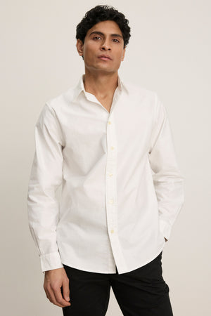 A man with short dark hair wearing the EMMETT BUTTON-DOWN SHIRT by Velvet by Graham & Spencer and black pants stands against a plain light background—a timeless wardrobe staple.