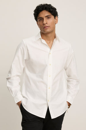 A man stands against a plain background, hands in his pockets, wearing the EMMETT BUTTON-DOWN SHIRT by Velvet by Graham & Spencer—a crisp white cotton oxford shirt—paired with black pants.