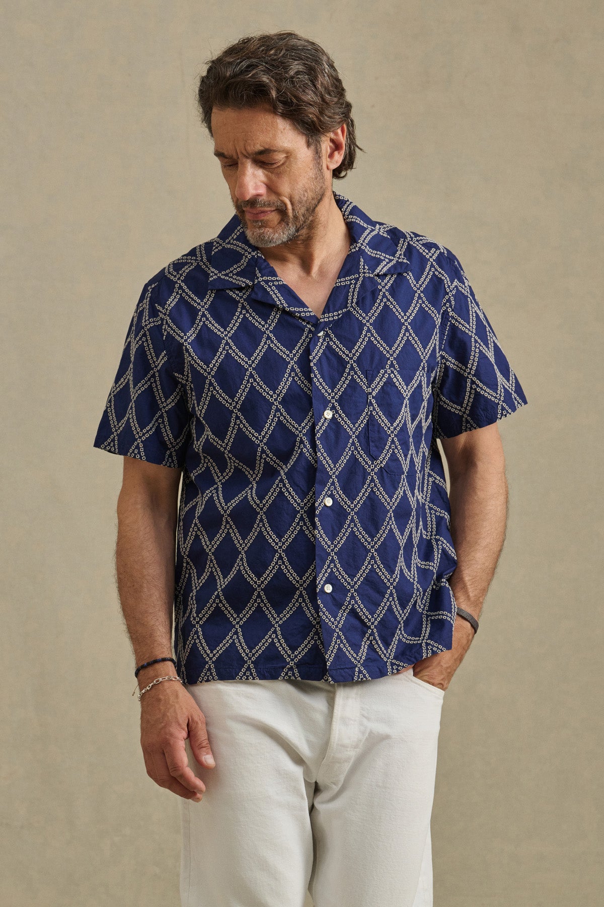 A man stands against a neutral background, looking down with one hand in his pocket, wearing the ESTEBAN SHIRT by Velvet by Graham & Spencer—a navy blue cotton shirt with white geometric embroidery—paired with white pants.-41626552697025