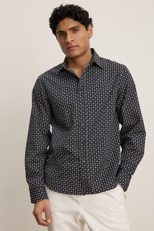 A man stands against a plain background wearing the LAWRENCE BUTTON-UP SHIRT by Velvet by Graham & Spencer, featuring a classic fit and dark pattern, paired with white pants for a sharp, tailored look.