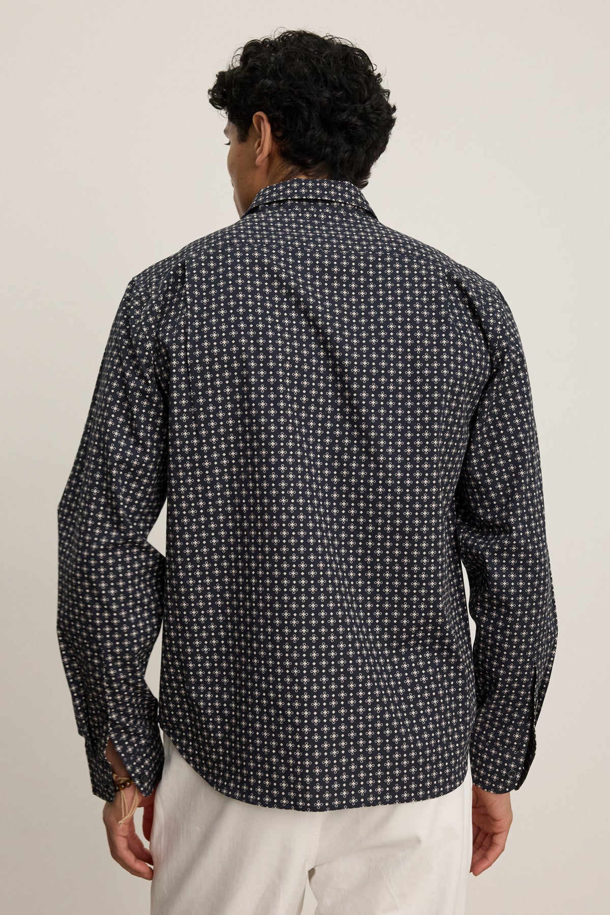 A person faces away, wearing Velvet by Graham & Spencer’s LAWRENCE BUTTON-UP SHIRT in a classic fit with a dark pattern and white pants, standing against a plain light background.