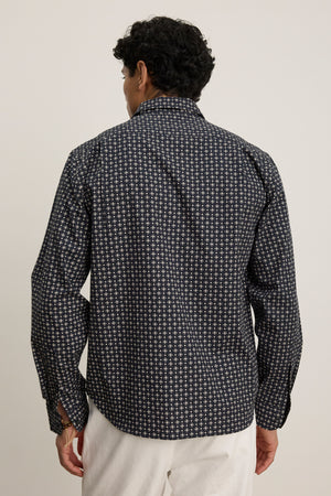A person faces away, wearing Velvet by Graham & Spencer’s LAWRENCE BUTTON-UP SHIRT in a classic fit with a dark pattern and white pants, standing against a plain light background.