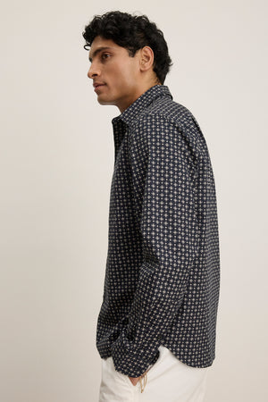 A man wearing the LAWRENCE BUTTON-UP SHIRT by Velvet by Graham & Spencer, styled in a classic fit with a dark pattern and paired with white pants, stands facing left against a plain background, showcasing his tailored look.