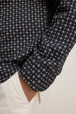 Close-up of a person wearing the LAWRENCE BUTTON-UP SHIRT by Velvet by Graham & Spencer, featuring a classic fit and navy blue pattern with white designs, paired with white pants and their hand in their pocket.