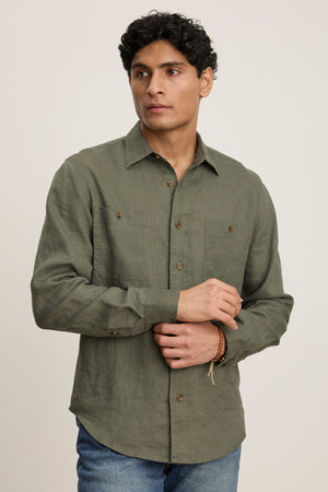 A man in a green LINDEN LINEN BUTTON-UP SHIRT by Velvet by Graham & Spencer and blue jeans stands against a plain light background, looking slightly to his left.