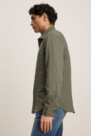 A person stands sideways against a plain light background wearing the LINDEN LINEN BUTTON-UP SHIRT by Velvet by Graham & Spencer in olive green, paired with blue jeans.