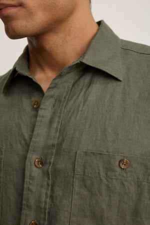 A close-up of a person wearing the LINDEN LINEN BUTTON-UP SHIRT by Velvet by Graham & Spencer in olive green, featuring a button front, two chest pockets, brown buttons, and an adjustable fit.