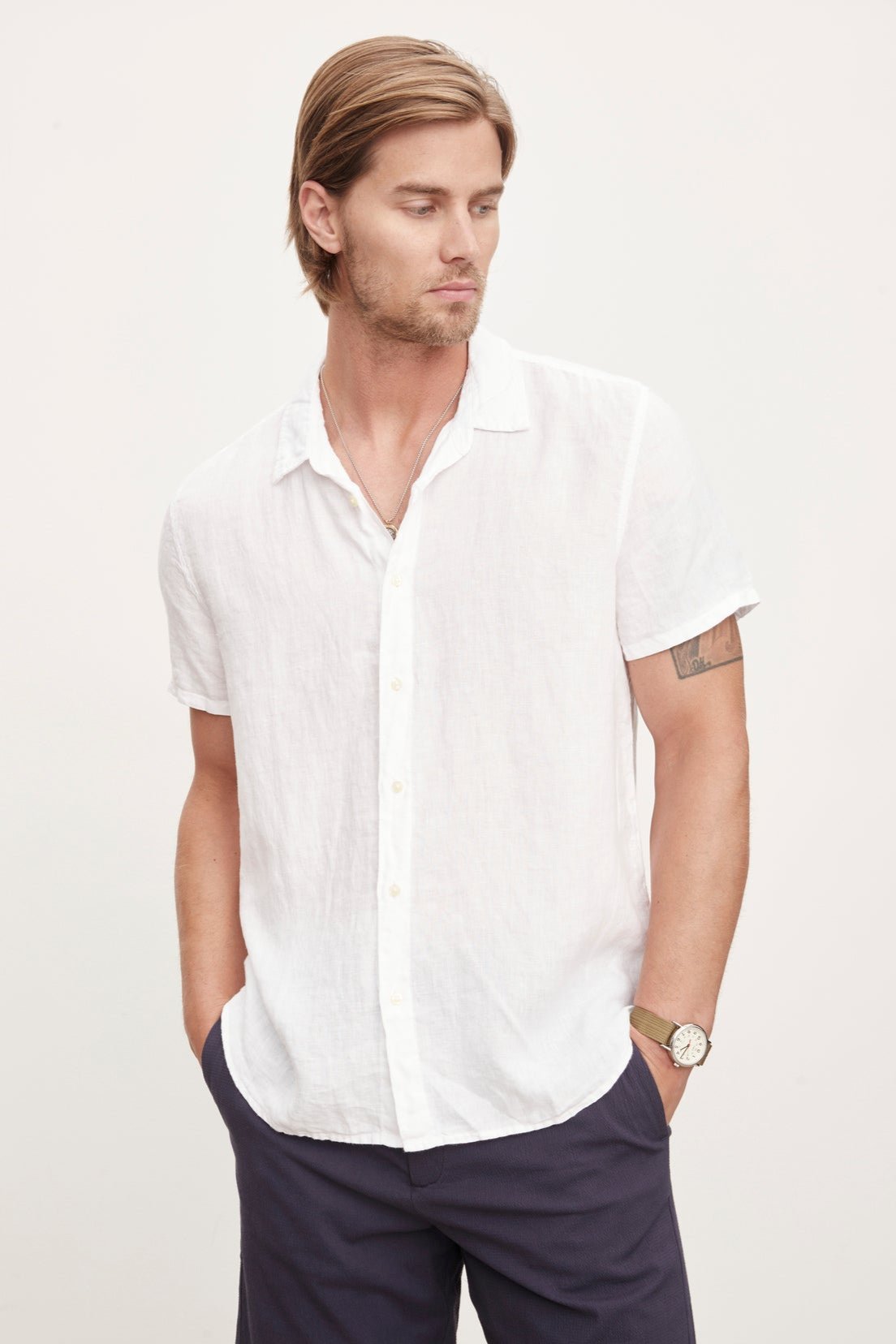 A man in a Velvet by Graham & Spencer MACKIE LINEN BUTTON-UP SHIRT and navy shorts, standing with one hand in his pocket and a watch on his left wrist, epitomizes the relaxed fit of a summer wardrobe.