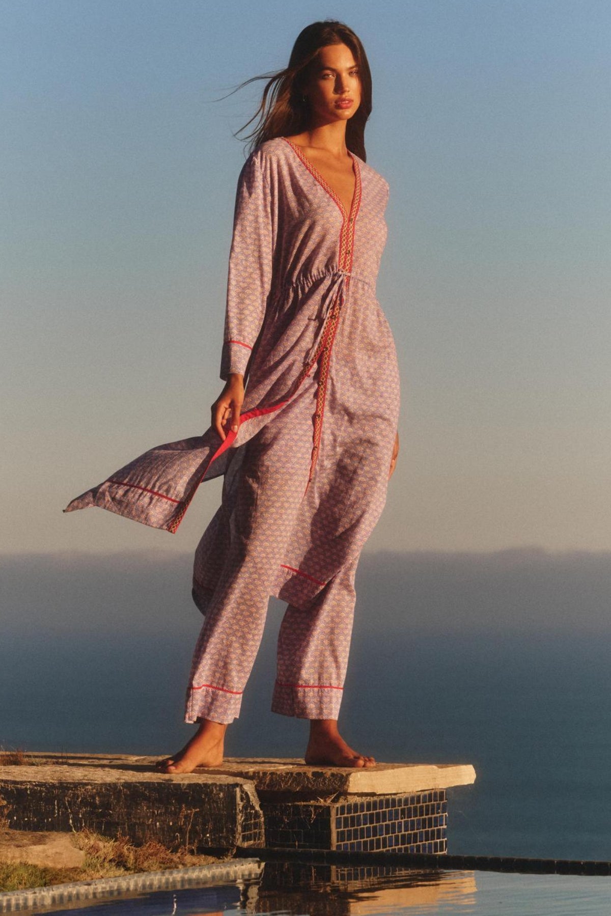 A woman stands barefoot by a pool, framed by ocean and sky, wearing the WRENLEY DRESS from Velvet by Graham & Spencer—a cotton voile midi dress with embroidered detailing.-40826601308353