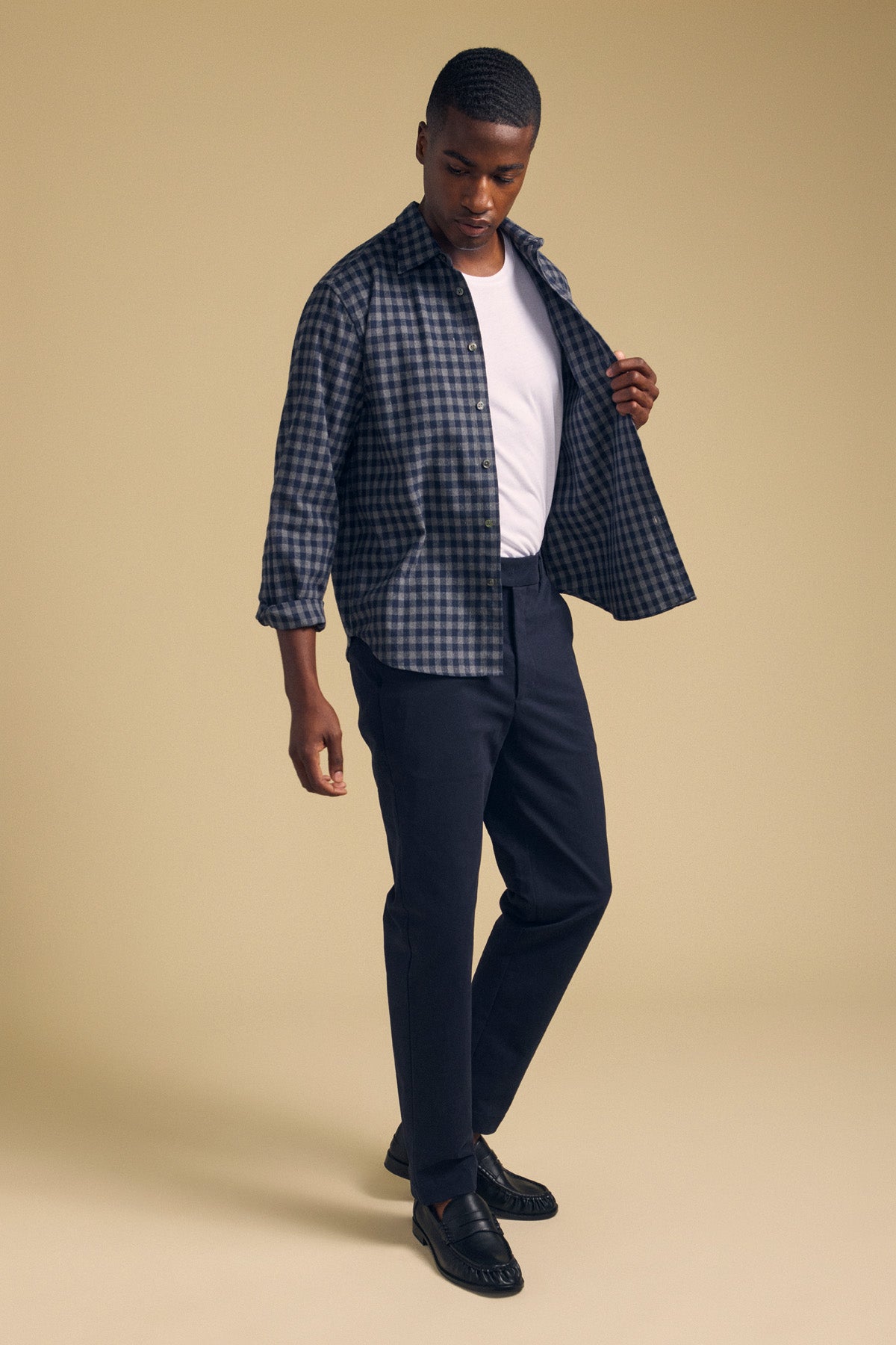 A man stands facing slightly downward against a beige background, wearing JUDE CHINO trousers by Velvet by Graham & Spencer, paired with a black and gray checkered shirt over a white t-shirt and black loafers.-41702027886785