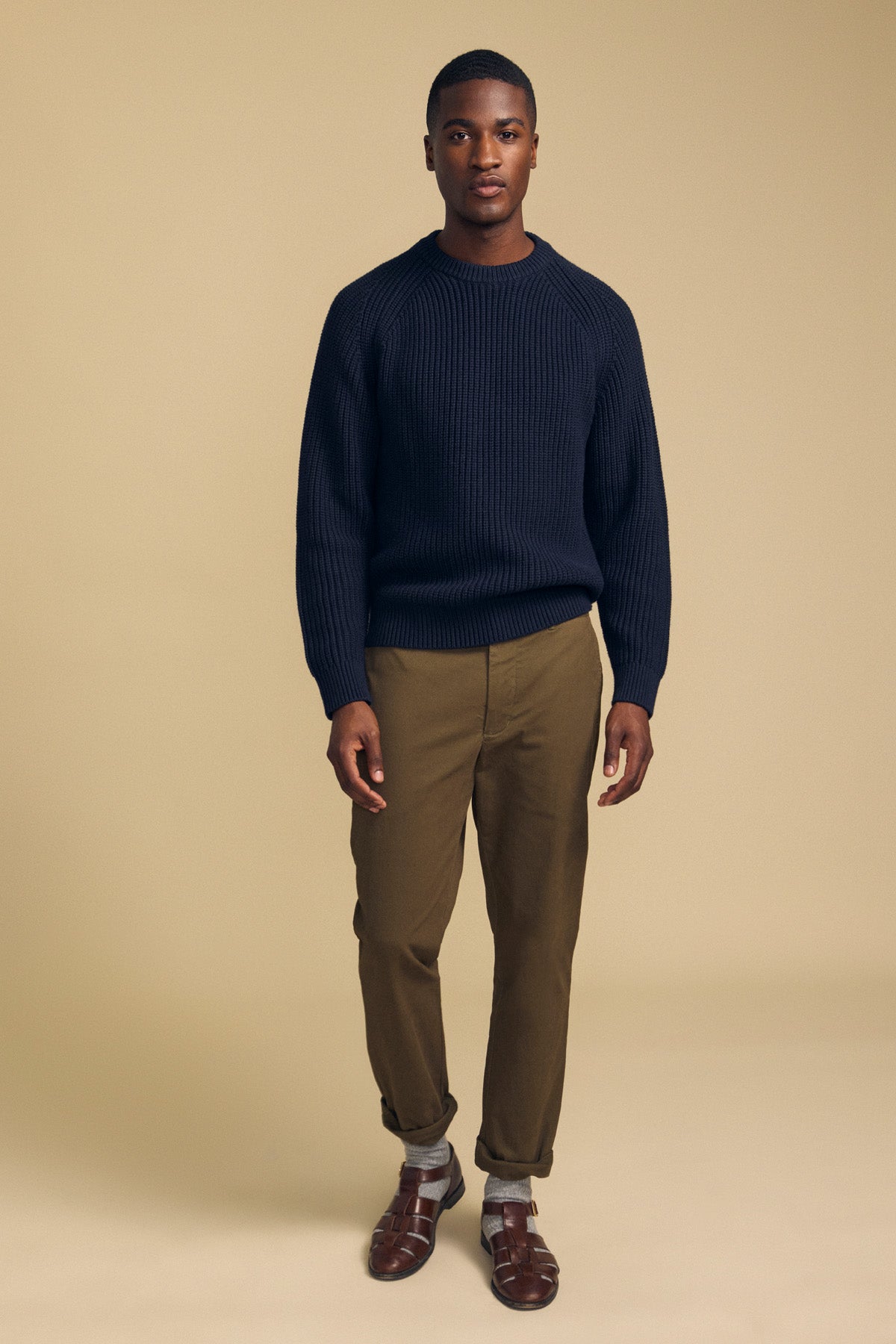 A man stands against a beige background wearing Velvet by Graham & Spencer LUIS CHINO trousers in olive, paired with a navy knit sweater, grey socks, and brown sandals. His cuffs are rolled, and he faces forward with arms relaxed at his sides.-41859720184001