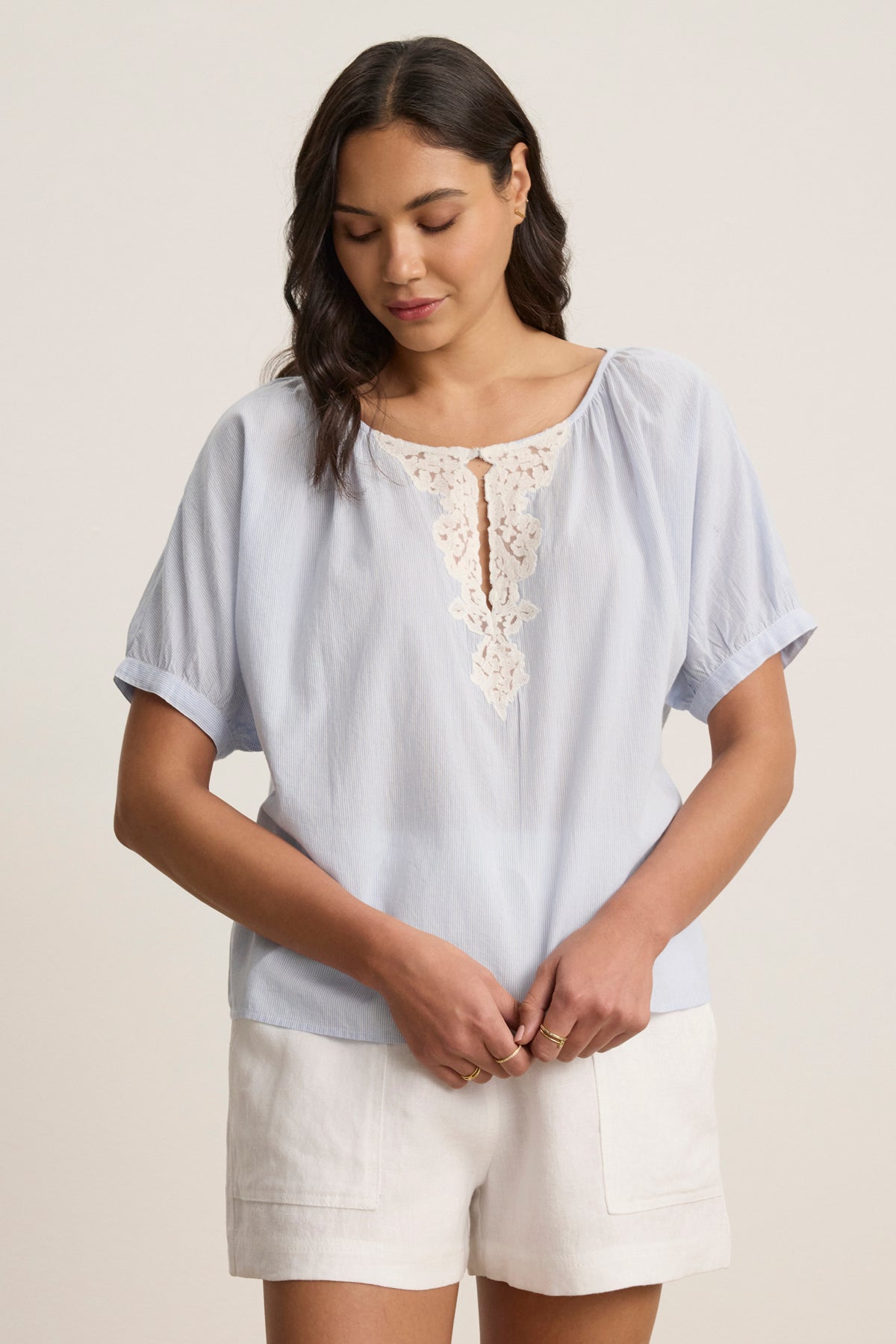 A woman stands wearing the PENELOPE BLOUSE by Velvet by Graham & Spencer, a light blue boatneck top with lace at the neckline, paired with white shorts. She looks down with her hands clasped.