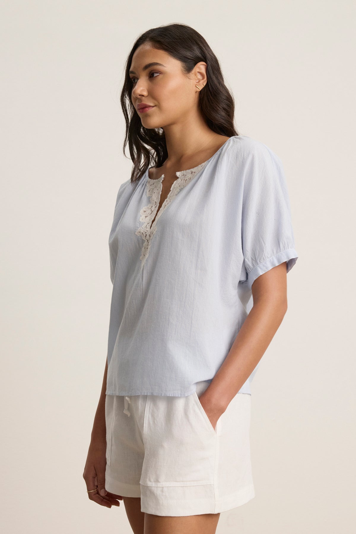A woman wears the PENELOPE BLOUSE by Velvet by Graham & Spencer—a light blue boatneck top with lace at the neckline—paired with white shorts, standing against a plain background.-39123353534657