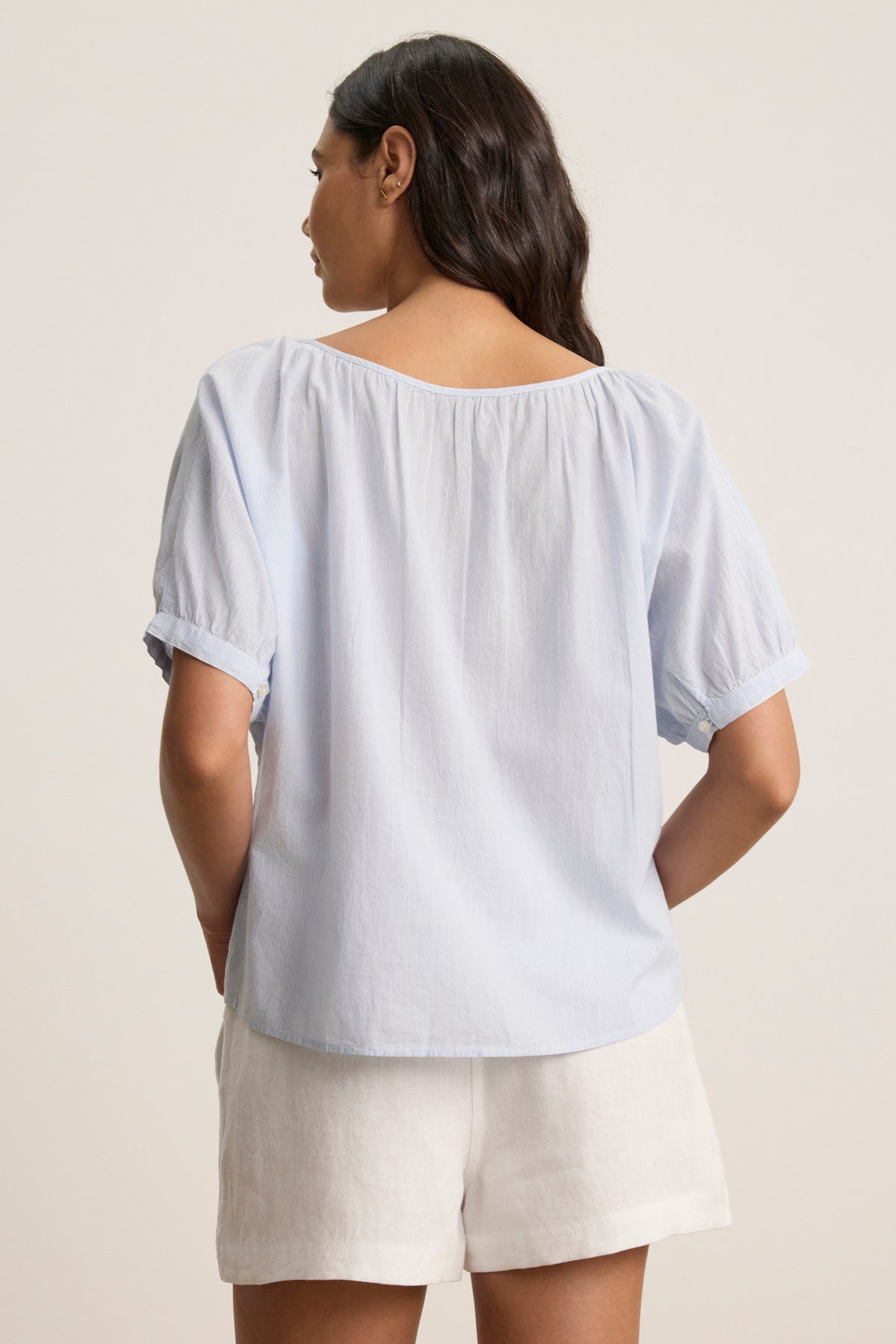 A woman, her hands in her pockets, faces away wearing the PENELOPE BLOUSE by Velvet by Graham & Spencer—a light blue boatneck top with lace details—paired with white shorts against a plain background.