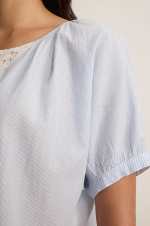 Close-up of a person wearing the PENELOPE BLOUSE by Velvet by Graham & Spencer, a light blue, short-sleeved, yarn-dyed top with delicate lace detailing at the neckline.