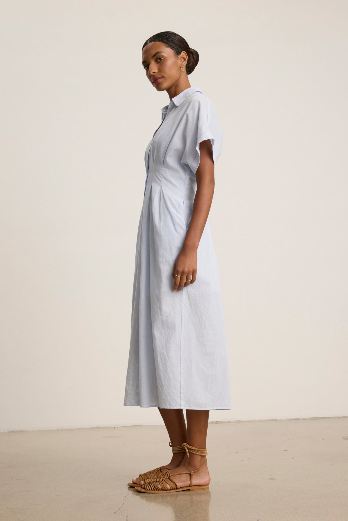 A woman stands sideways, highlighting the timeless elegance of the Velvet by Graham & Spencer RILEY DRESS—a light blue, short-sleeve, collared midi—paired with brown woven sandals against a plain background.-39123354190017