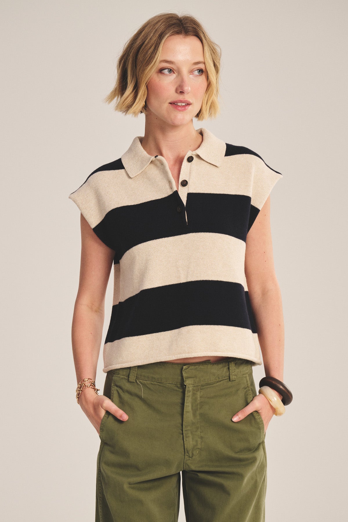 A woman wears the ZOELLA SWEATER POLO by Velvet by Graham & Spencer—a cream and black striped, cropped sweater polo—tucked into olive green pants, with her hands in her pockets.-40910161543361
