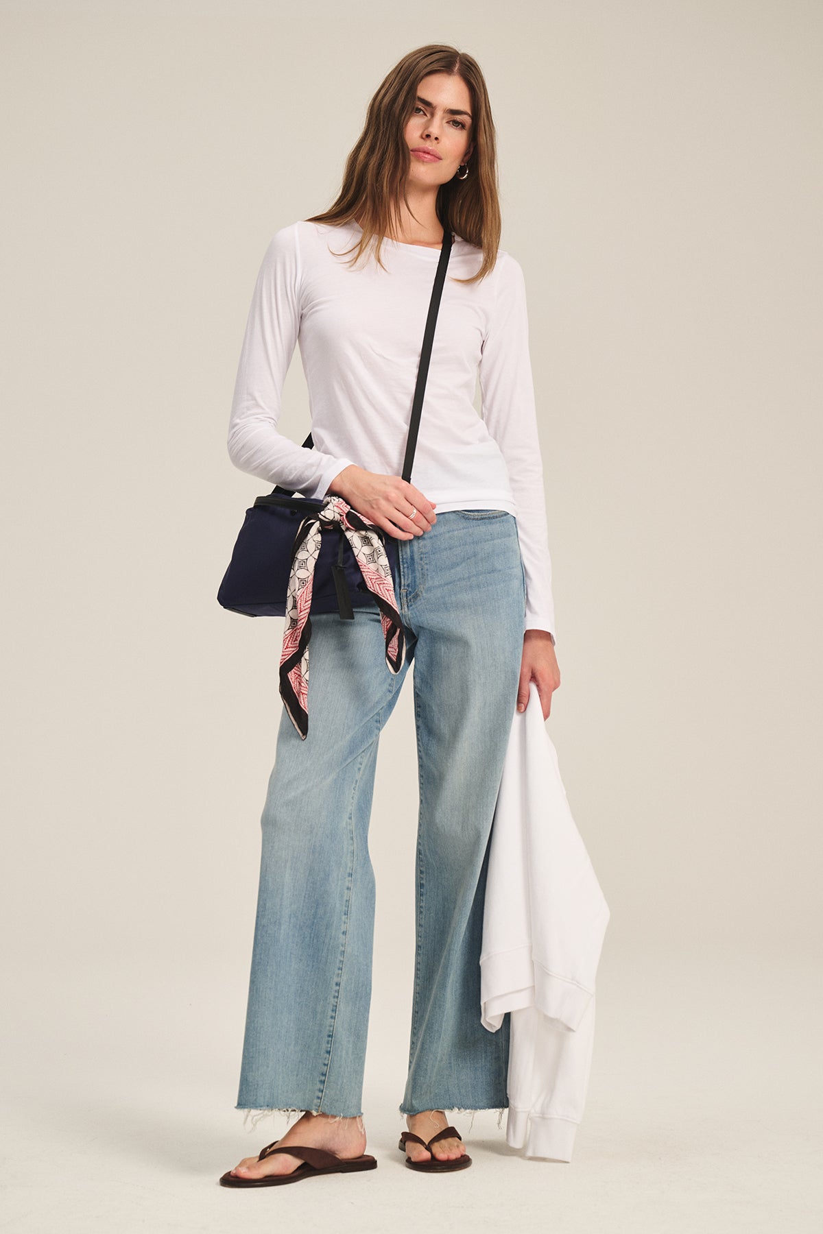 A person stands against a plain background wearing the Velvet by Graham & Spencer ZOFINA TEE—a fitted long sleeve crewneck—paired with wide-leg jeans, sandals, a crossbody bag, and holding a white jacket.