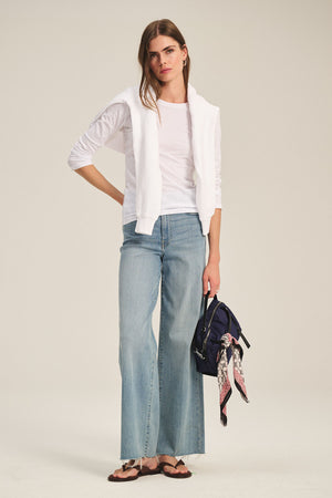 A person stands against a plain background wearing a white long-sleeve shirt, sweater over the shoulders, wide-leg jeans, sandals, and carrying the Velvet by Graham & Spencer NYLON CROSSBODY BAG with a patterned scarf tied to its strap.
