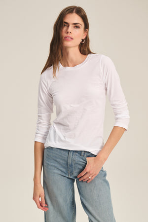 A woman stands against a plain, light background wearing the ZOFINA TEE by Velvet by Graham & Spencer—an ultra-soft, fitted long sleeve crewneck—paired with light blue jeans.