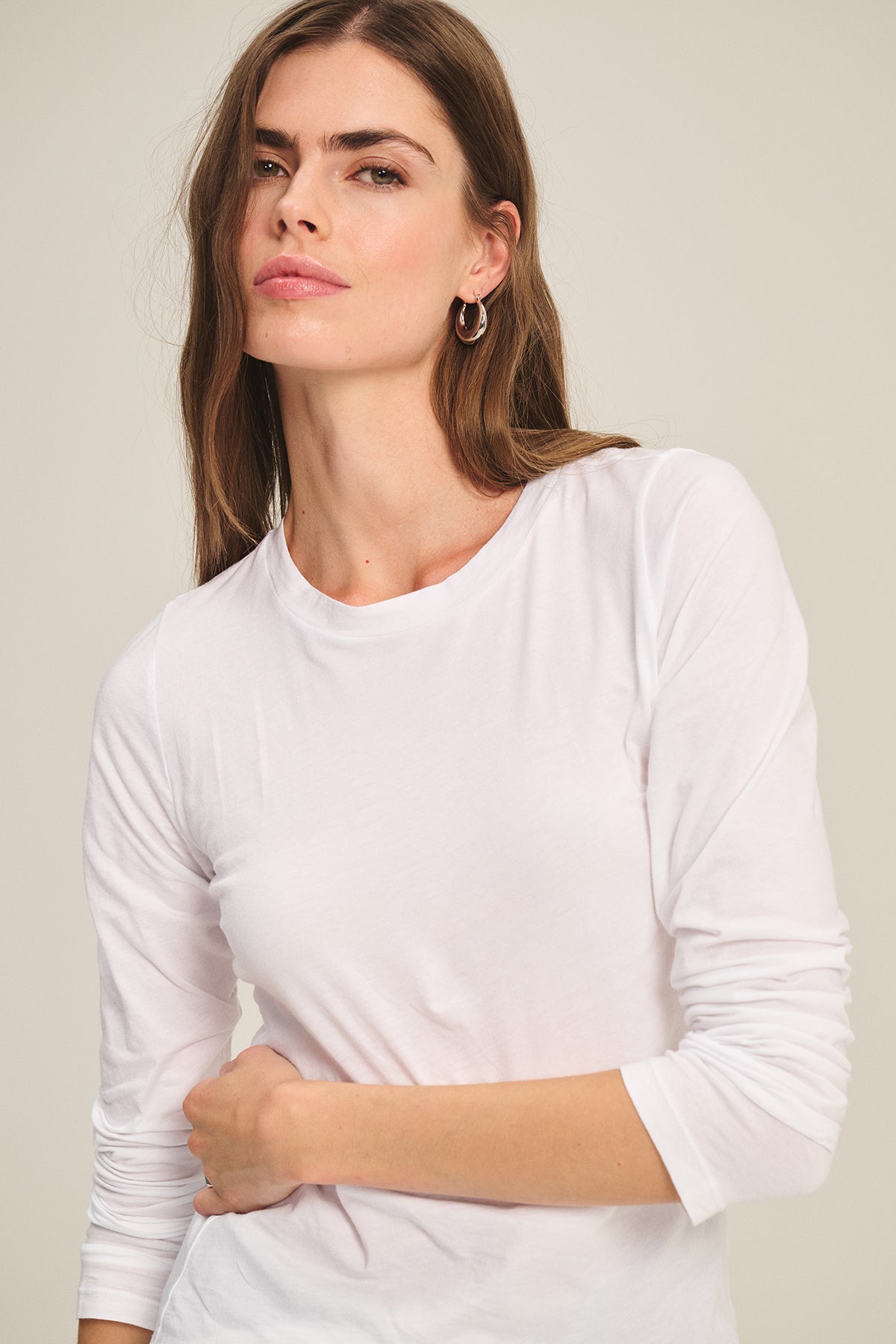 A woman with long brown hair wears the ZOFINA TEE by Velvet by Graham & Spencer, a fitted long sleeve crafted from ultra-soft fabric, standing against a neutral background and gazing slightly to the side.-39601037279425