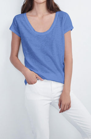 A woman is standing against a white background, wearing the KIRA TEE from Velvet by Graham & Spencer made from cotton slub fabric and white pants, perfect for everyday wear. Her left hand is in the pocket of her pants.