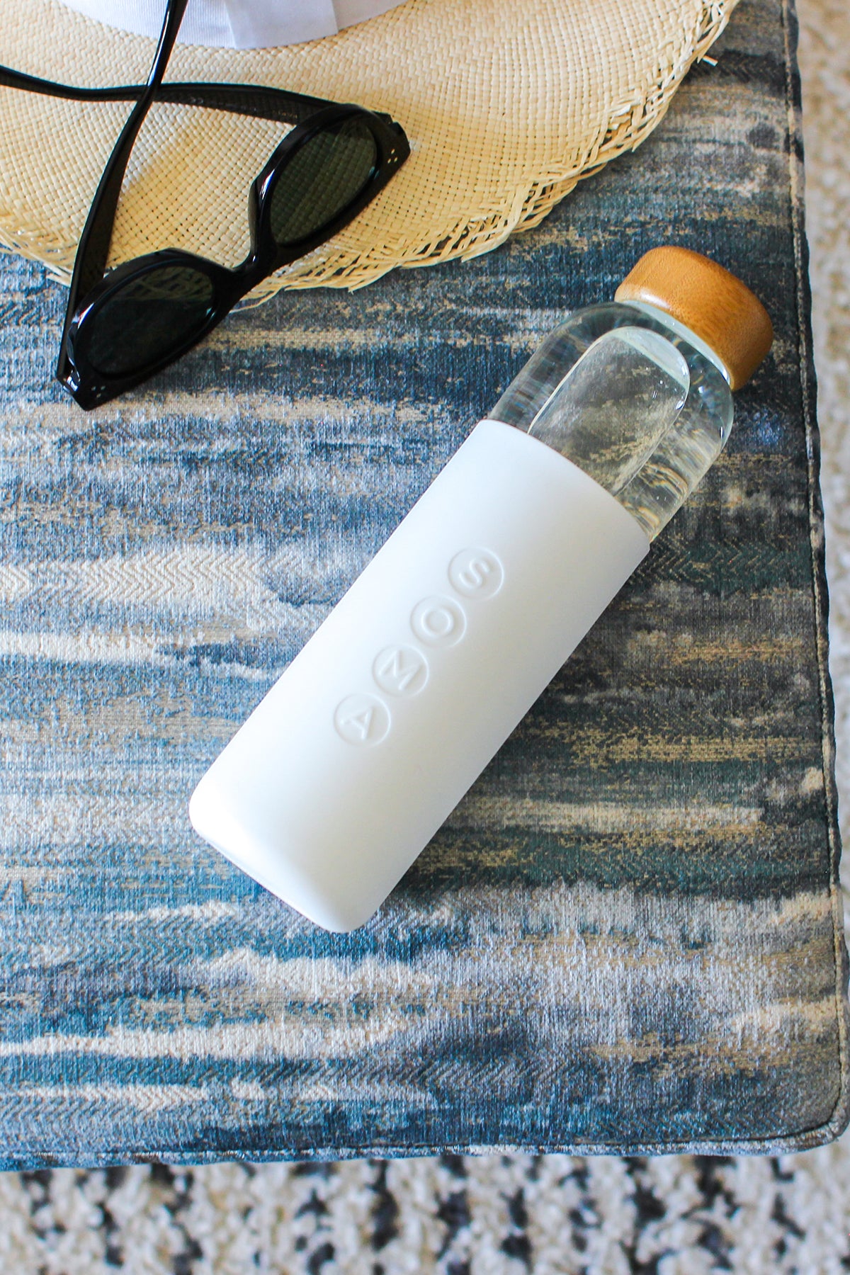 The GLASS WATER BOTTLE BY SOMA by Soma, with an eco-friendly bamboo cap and white silicone sleeve, rests on a blue textured surface next to black sunglasses and a straw hat—ideal for anyone seeking a sustainable water bottle.-8128185434193