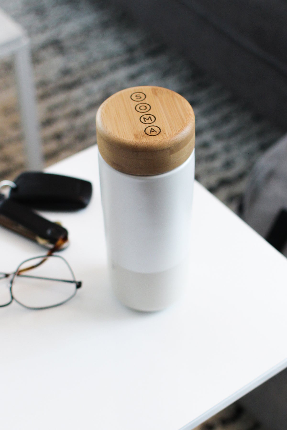 The Soma INSULATED CERAMIC MUG, featuring a sleek white design and a leak-proof bamboo lid, rests on a white table beside eyeglasses and keys.-8128159318097