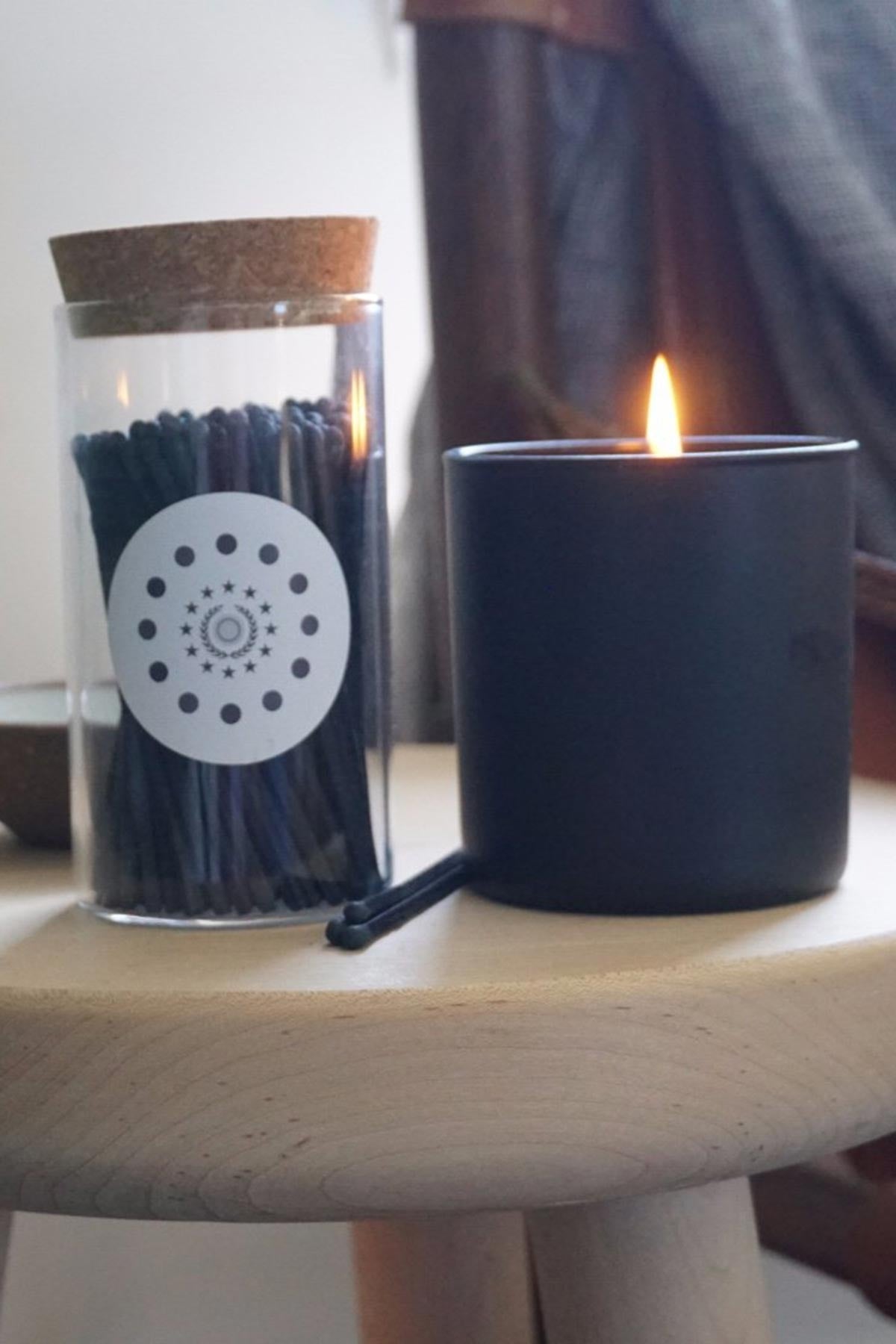 A black candle sits on wood beside Frederick and Mae’s MATCHES BY FREDRICKS AND MAE—black matches in a glass jar sealed with a cork lid.-555779686481