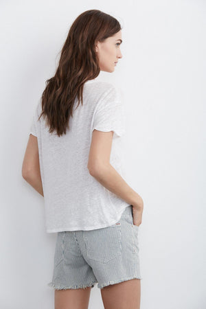 A woman with long brown hair stands against a white background, wearing a white t-shirt and the Cindy Frayed Hem Boyfriend Shorts by Velvet by Graham & Spencer. She's facing away, with her left hand in her shorts pocket, showcasing the relaxed silhouette and frayed hems.