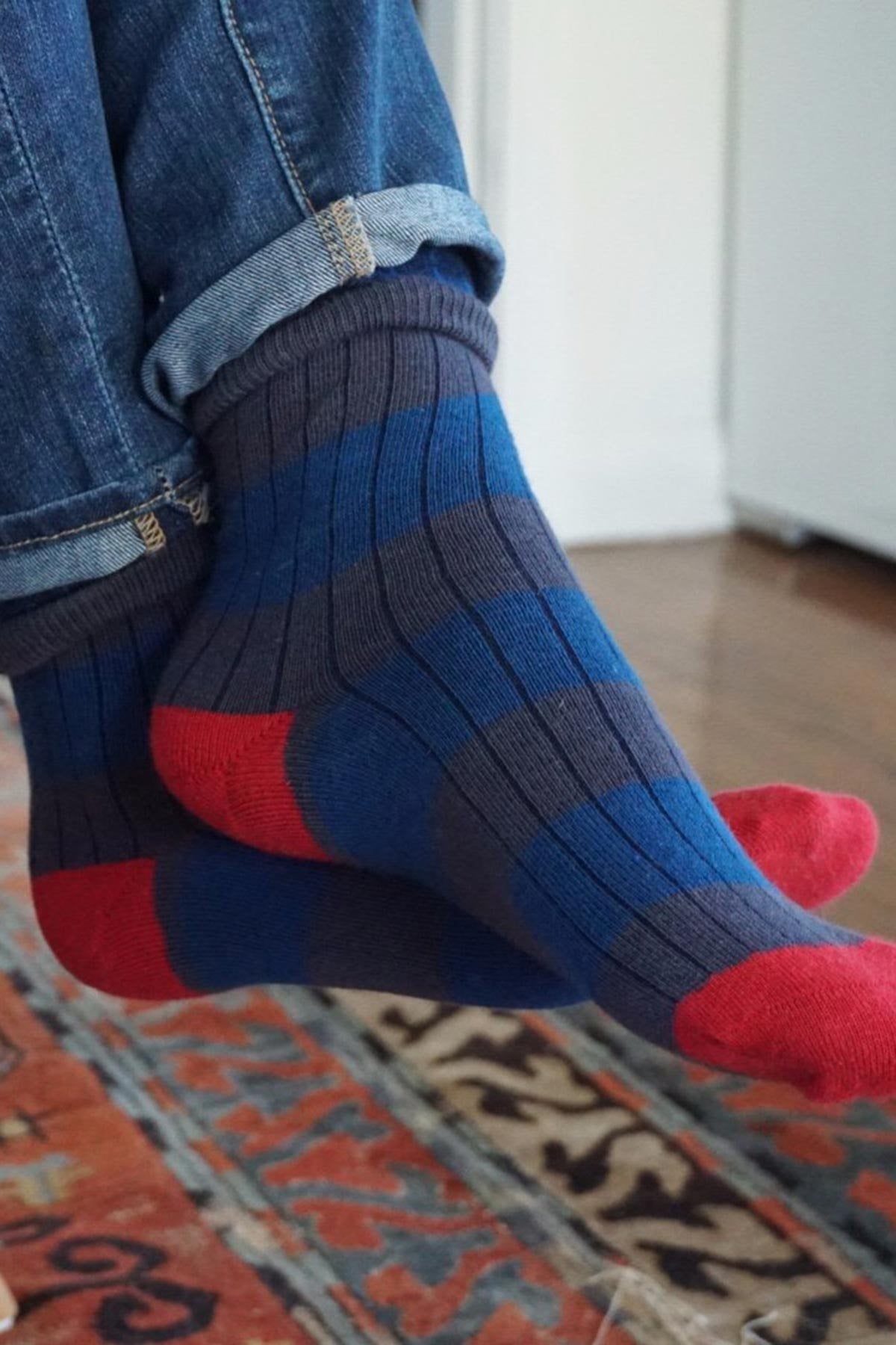 A person in rolled-up blue jeans and Little River's STRIPED RIB CREW SOCK by Little River Sock Mill sits cross-legged on a patterned rug, showing off their blue-and-red striped organic cotton socks.-668331671633