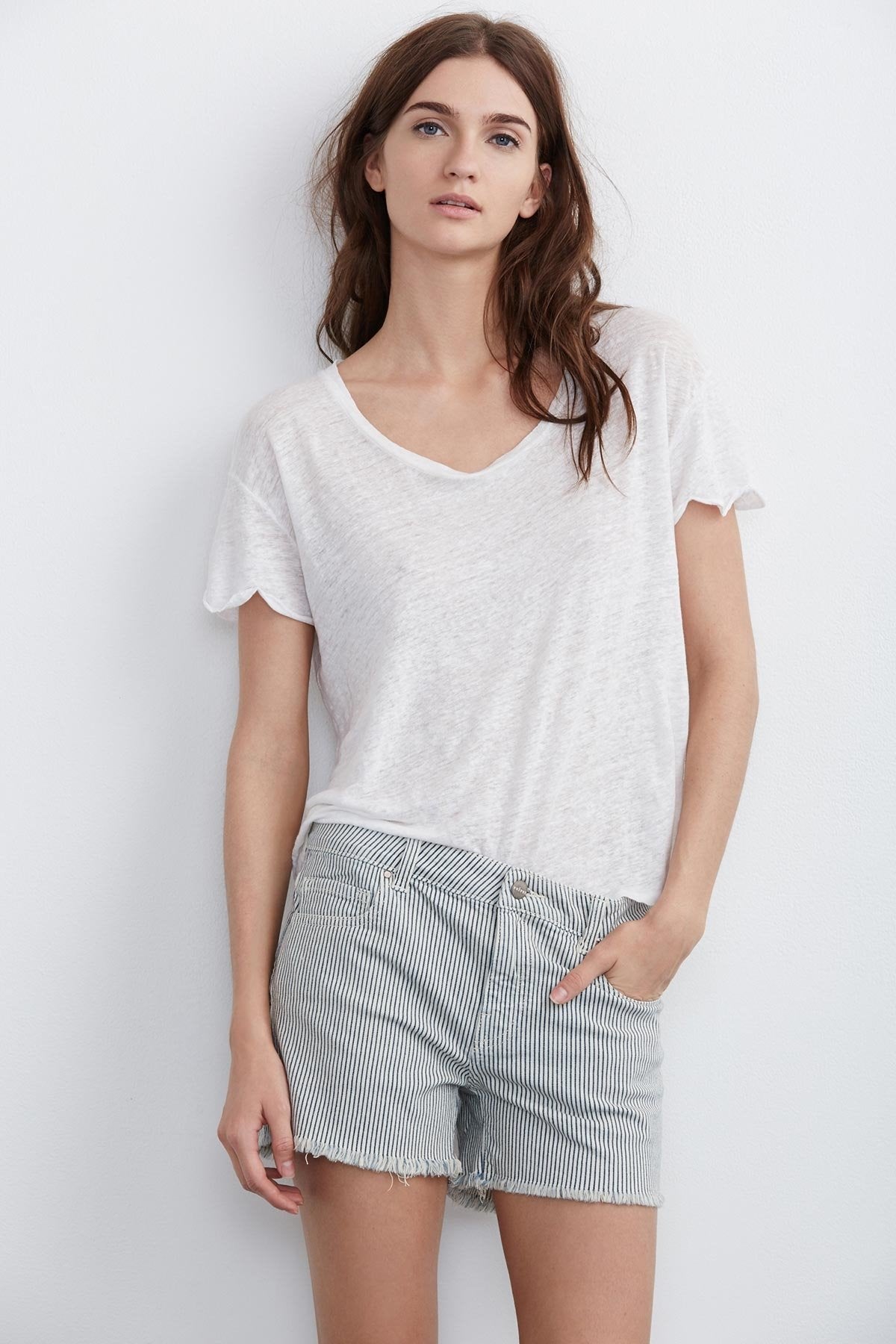 A woman with long, dark hair wearing a white t-shirt and the Cindy frayed hem boyfriend shorts by Velvet by Graham & Spencer stands against a white background with one hand in her pocket, exuding a relaxed silhouette.