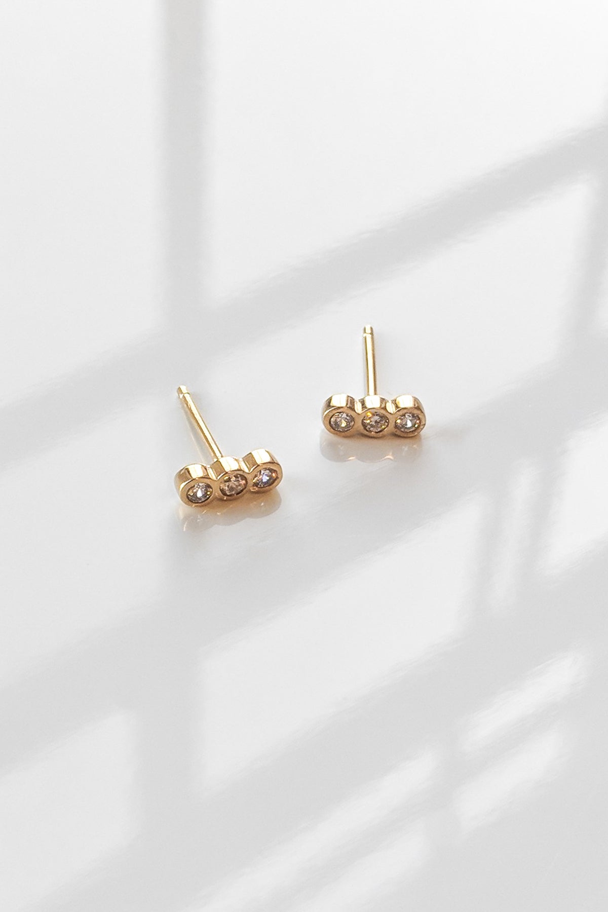 The ILER STUD EARRINGS BY THATCH, crafted by Thatch, are gold vermeil with three gemstones each, elegantly shown on a reflective white surface with intricate shadows. These semi-precious stone studs exude elegance and charm.