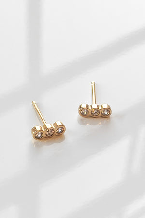 The ILER STUD EARRINGS BY THATCH, featuring three small round diamonds each, placed on a reflective white surface, highlight the charm of Thatch's handcrafted jewelry.
