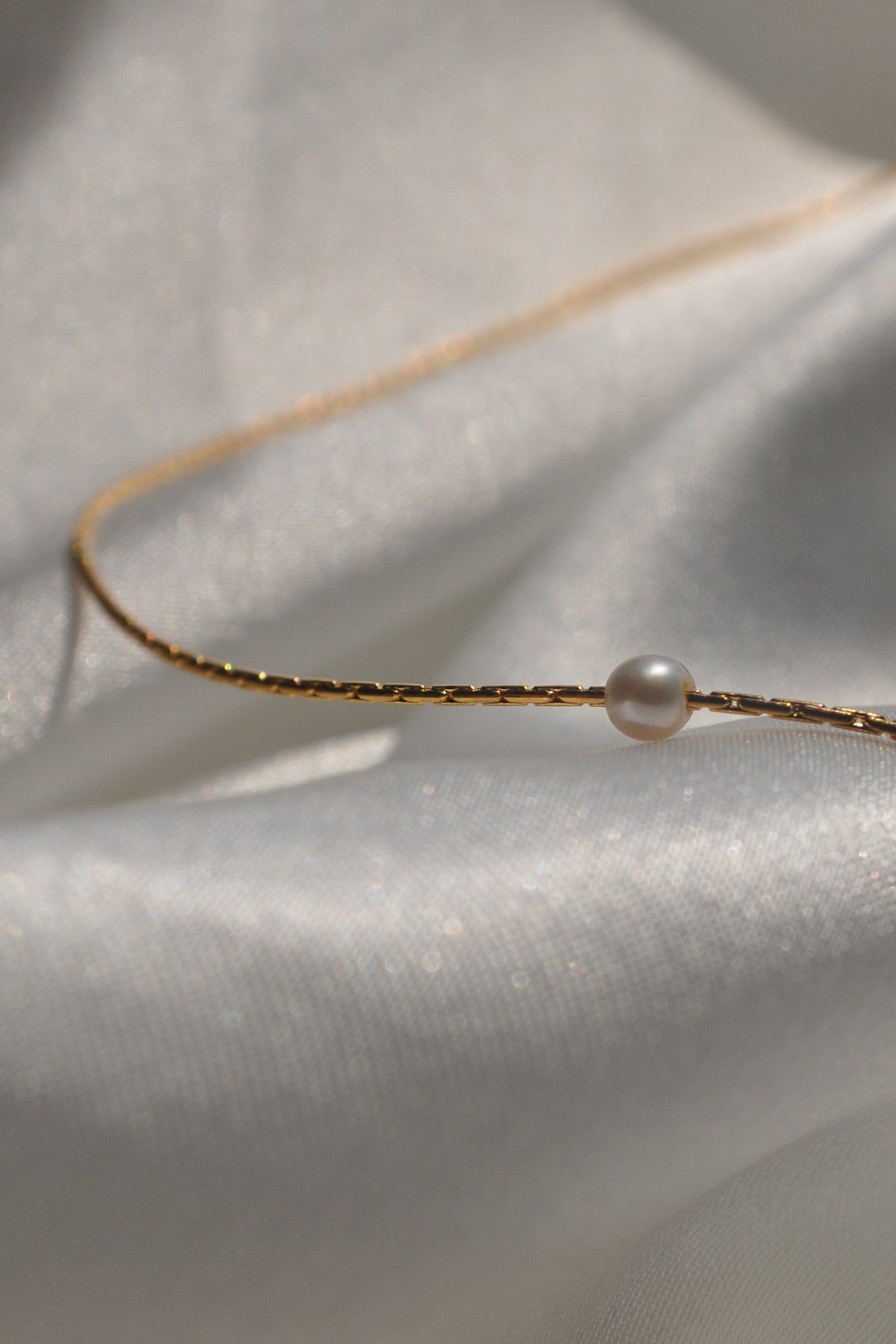 The MINA NECKLACE BY THATCH, a refined 14K gold snake chain adorned with a single freshwater pearl, is elegantly placed on shimmering fabric, highlighting the Thatch jewelry collection's sophistication.