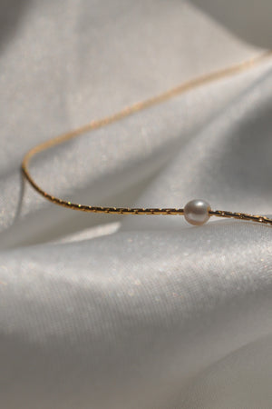 The MINA NECKLACE BY THATCH, a refined 14K gold snake chain adorned with a single freshwater pearl, is elegantly placed on shimmering fabric, highlighting the Thatch jewelry collection's sophistication.