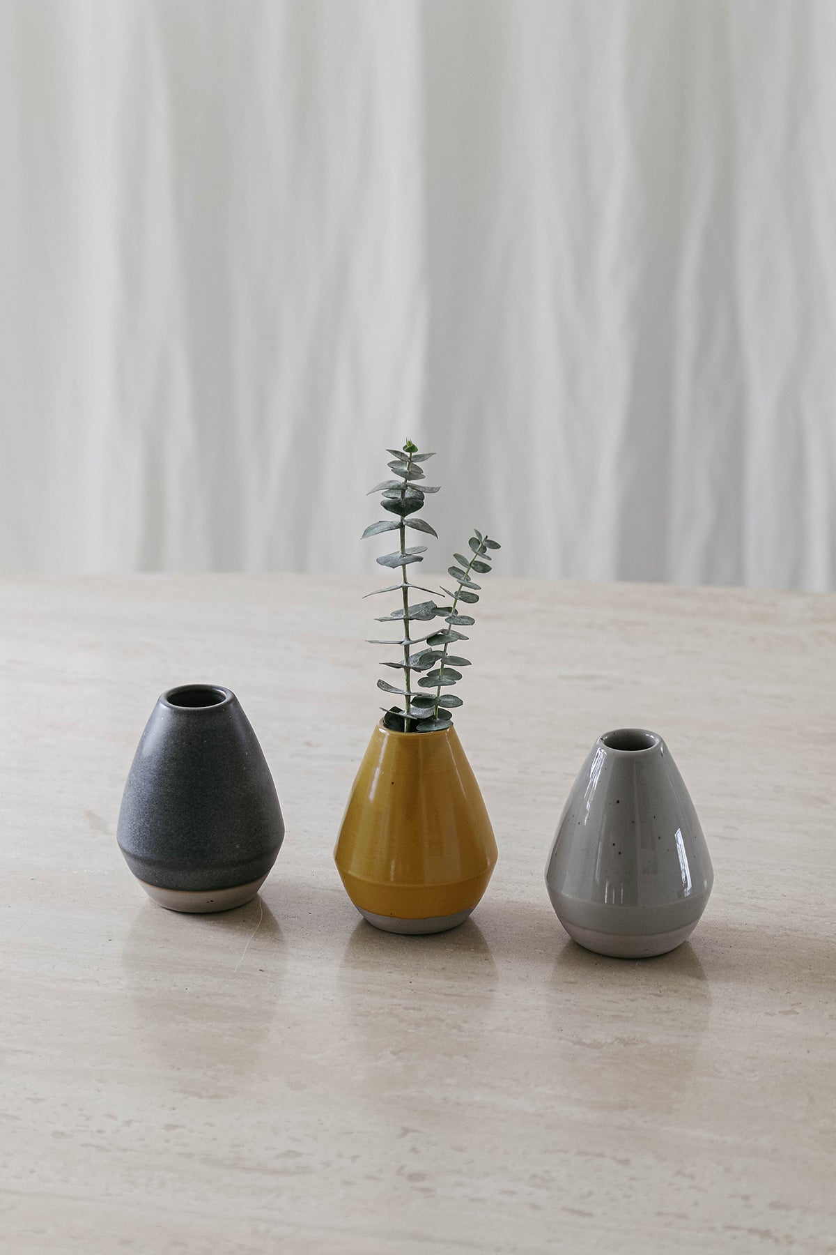 Three petite Jenny Graham Home ceramic bud vases with single ranunculus plants in ceramic vases on a table.-25629356916929