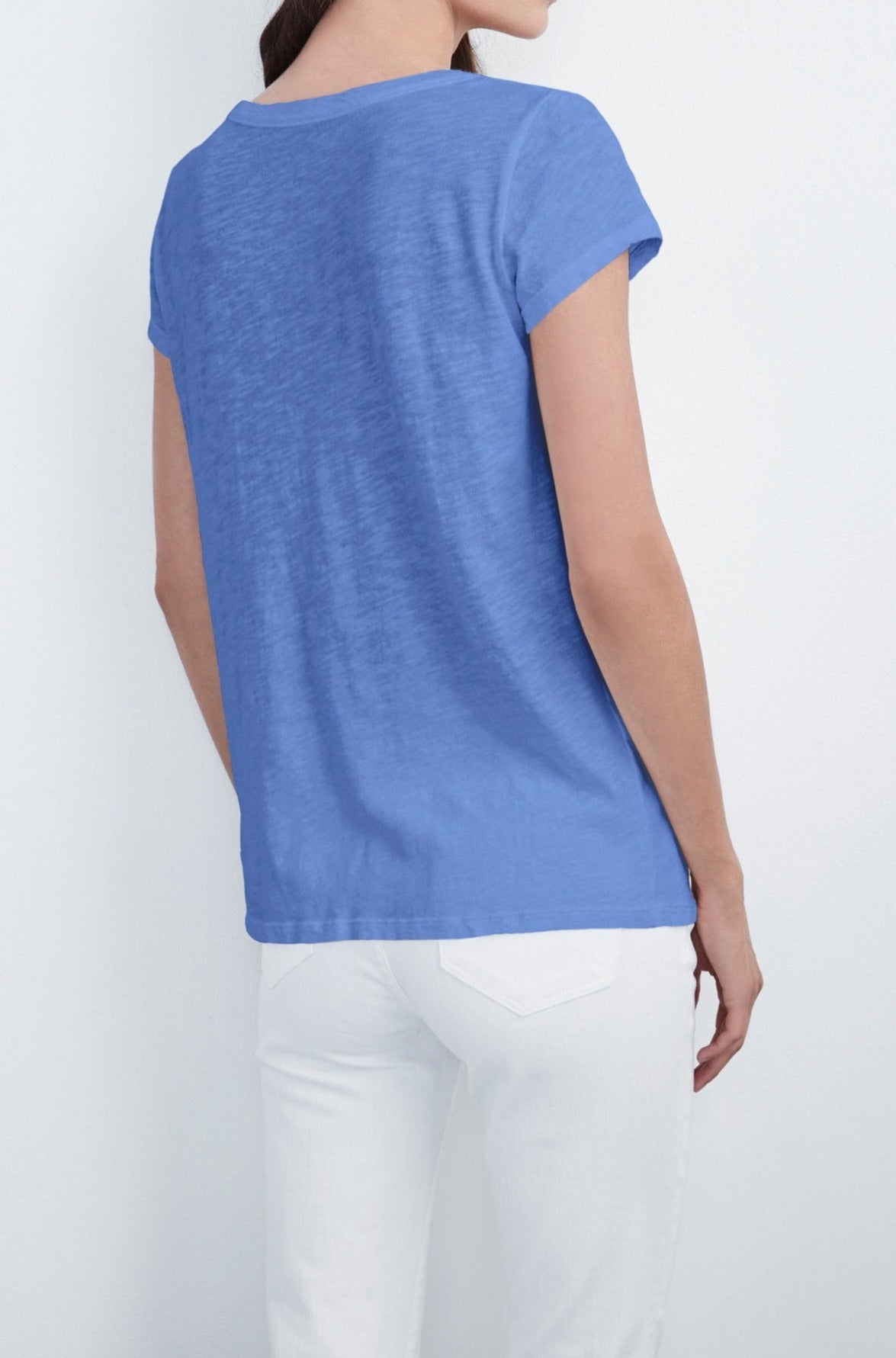 A person is standing with their back to the camera, wearing a blue short-sleeve KIRA TEE by Velvet by Graham & Spencer with a scoop neckline and white pants, effortlessly showcasing everyday wear.-555721785425