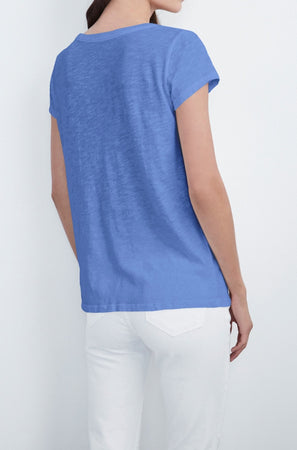 A person is standing with their back to the camera, wearing a blue short-sleeve KIRA TEE by Velvet by Graham & Spencer with a scoop neckline and white pants, effortlessly showcasing everyday wear.