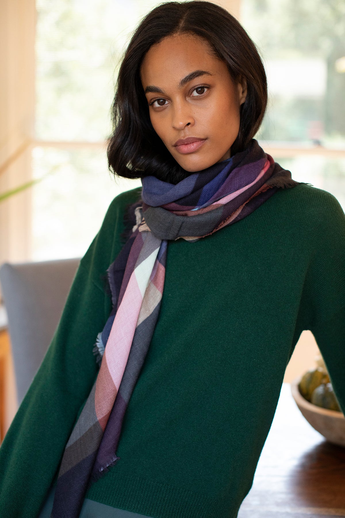 A woman with shoulder-length dark hair, wearing a green sweater and the PLAID WOOL BLEND SCARF BY EPICE from Epice, stands indoors facing the camera.
