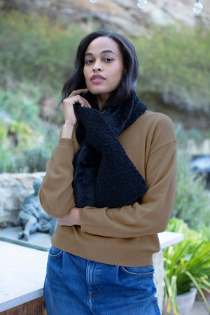 A woman stands outdoors amid greenery and stone, wearing a brown sweater, blue jeans, and the Velvet by Graham & Spencer REVERSIBLE SHERPA SCARF.