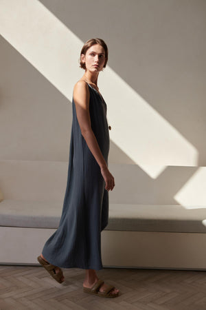 A woman in a blue cotton gauze Carrillo Dress by Velvet by Jenny Graham stands in a sunlit room with diagonal light patterns on the wall, looking thoughtfully to the side.