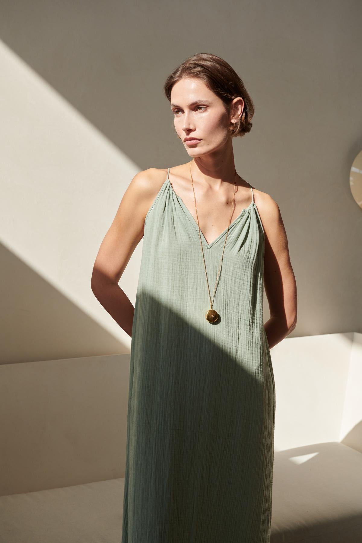 A woman in a Velvet by Jenny Graham CARRILLO DRESS stands in a sunlit room with shadows cast by a window, looking thoughtfully to the side.-26293219786945